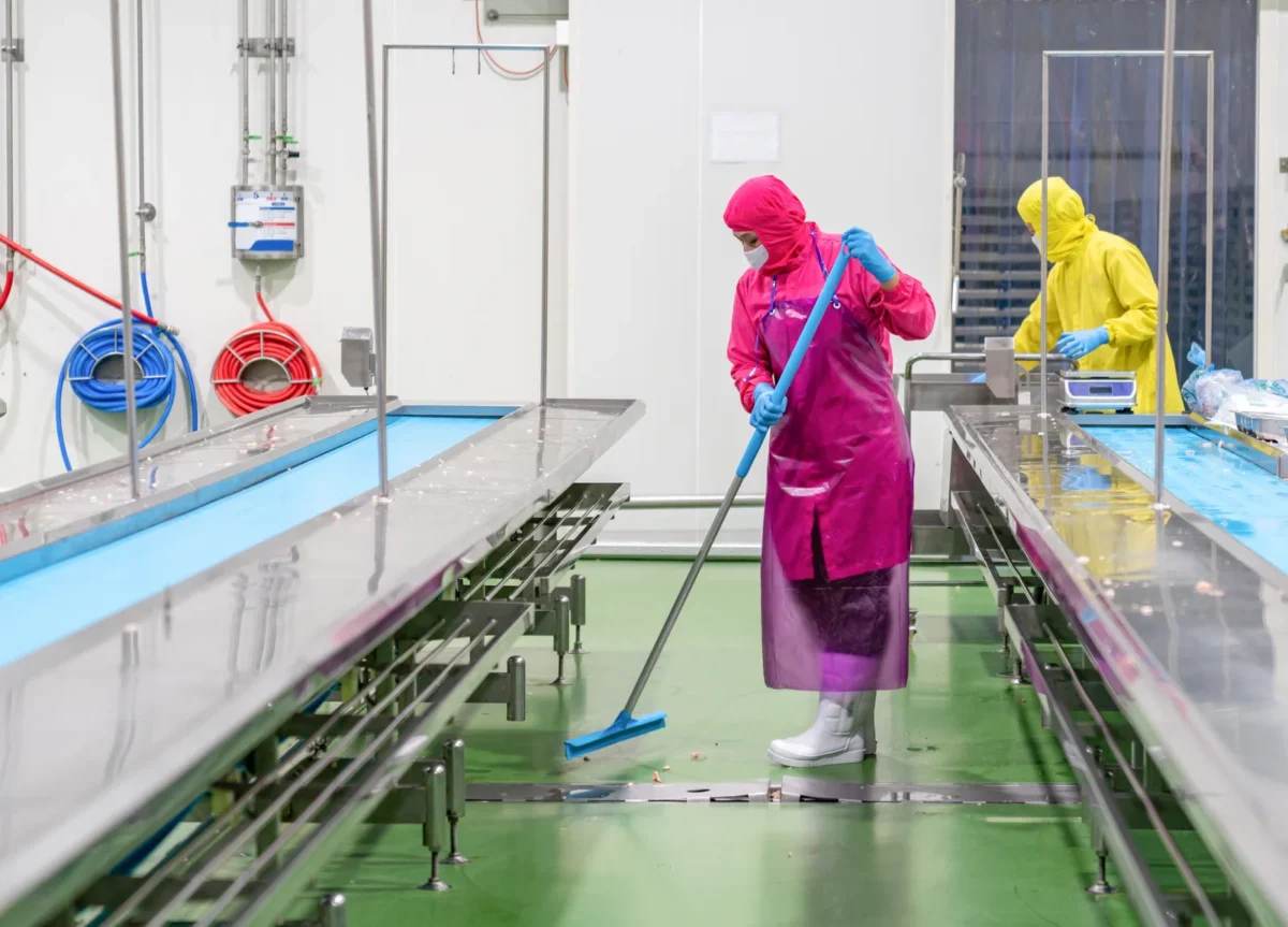 Workers washing a food-production floor beside processing conveyors